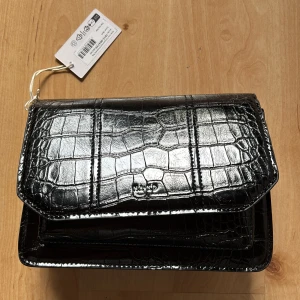 Svart axelväska i krokopräglad syntet från NA-KD - Säljer en svart axelväska från NA-KD i krokopräglad syntet. Väskan har ett lock med magnetknäppning, innerfack med dragkedja och bred avtagbar axelrem i textil. Perfekt för att bära det viktigaste med stil.