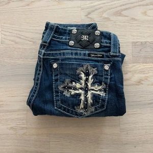 Miss Me Jeans med Svarta Detaljer  - Säljer ett par mörkblå jeans från Miss Me med snyggt broderad bakficka i silver och svart. Jeansen har dekorativa knappar och detaljer, samt klassisk femficksdesign. Bootcut-modell med låg midja och slitningar för en cool look.Längd 99 midja 33-35cm benöppning 20cm