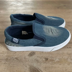 Jeansblå slip-on sneakers från Pop Café - Säljer ett par jeansblå slip-on sneakers från Pop Café med vit platt sula och slitna detaljer framtill. Skorna har elastiska sidor för enkel påtagning och är tillverkade i jeansmaterial. Perfekta för en avslappnad stil.