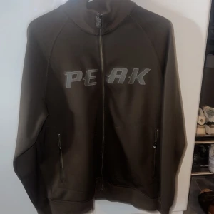 Brun zipjacka från Peak - Mörkbrun jacka med dragkedja från Peak. Jackan har hög krage, två fickor med dragkedja och tryckt logga på bröstet. Perfekt för en sportig och avslappnad stil.