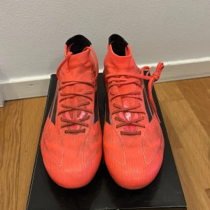 Adidas F50 Elite mid AG W - Säljer ett par röda Adidas f50 elite fotbollsskor med svarta ränder och snörning. Skorna har hög krage för extra stöd och platt dobbsula för bra grepp på planen. Perfekta för dig som vill sticka ut på fotbollsplanen. Skorna nypris 2999kr