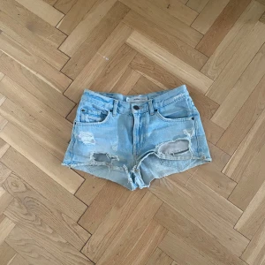 Ljusblå jeansshorts med slitningar - Säljer ett par ljusblå jeansshorts som är mid/low waist. Klassisk femficksmodell med knapp och dragkedja. Perfekta för varma dagar och en avslappnad stil.