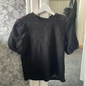 Svart kortärmar med puff - Säljer en svart blus från Object med vackert spetsmönster och puffiga korta ärmar. Blusen har rund halsringning och är tillverkad i ett genombrutet tyg som ger en elegant känsla. Perfekt att styla till olika outfits!