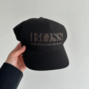 Svart keps med glittrig text - Snygg svart keps med glittrig text framtill som stavar 'BOSSY'. Perfekt accessoar för att lyfta din outfit med lite extra attityd. Justerbar passform baktill.