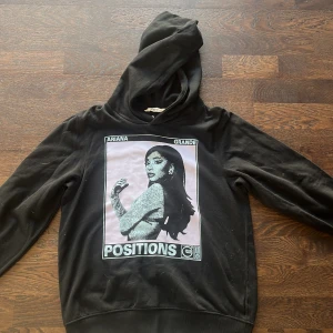 Svart hoodie Ariana Grande Positions - Svart hoodie med stor tryckt bild av Ariana Grande och texten 'POSITIONS' på framsidan. Tröjan har huva och långa ärmar. Perfekt för fans av Ariana Grande.