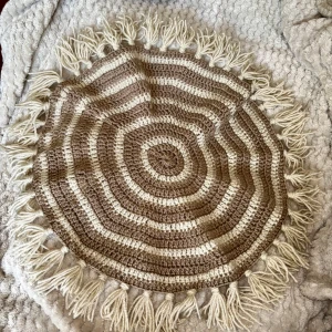 Handvirkad rund duk i naturtoner – unikt hantverk - Charmig och mjuk rund duk, handvirkad i varma naturfärger (brunt och beige) med dekorativa fransar runt kanten. Perfekt som inredningsdetalj på soffbord, byrå eller fönsterbräda. 	•	Diameter: ca 35 cm 	•	Material: 100 % akrylgarn (4 nystan) 	•	Färg: Beige och brunt i ränder 	•	Handgjord med omsorg under cirka en veckas arbete  Passar dig som gillar det personliga och unika i hemmet, eller som en fin present till någon du tycker om.  Pris: 250 kr