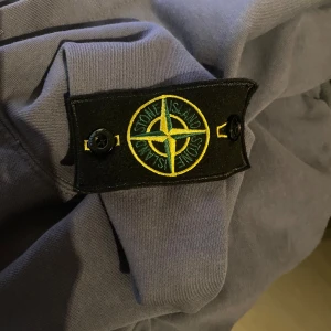 Blå huvtröja från Stone Island - Säljer en snygg blå huvtröja från Stone Island med dragkedja och huva. Tröjan har en ikonisk logga på ärmen och är perfekt för en casual look. Den har långa ärmar och ribbade muddar för extra komfort. Knappt använd 