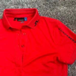 Röd pikétröja från J.Lindeberg - Säljer en snygg röd pikétröja från J.Lindeberg med klassisk krage och knappar framtill. Tröjan har logga på kragen och ärmen samt en liten logga i nacken. Perfekt för en sportig och stilren look. Är storlek small, men känns mer som XS.