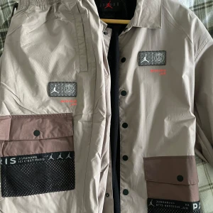Beige limited edition vindjacka samt byxor från Jordan x Paris Saint-Germain samt  - Säljer en beige vindjacka från Jordan x Paris Saint-Germain med svarta och bruna detaljer. Jackan har tryckknappar, dragkedja och flera fickor framtill. Stora tryck med Paris och Jordan-logga på ryggen. Perfekt för streetwear.