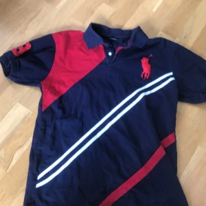 Mörkblå pikétröja från Polo Ralph Lauren - Säljer en mörkblå pikétröja från Polo Ralph Lauren i storlek XL. Tröjan har röda och vita detaljer, broderad logga på bröstet och tryck på ärmen. Klassisk krage och korta ärmar. Perfekt för en sportig och stilren look.