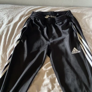 Svarta träningsbyxor från Adidas med vita ränder - Svarta träningsbyxor från Adidas med klassiska vita ränder längs sidorna och broderad logga på benet. Byxorna har resår i midjan och är tillverkade i ett syntetmaterial som passar perfekt för sport och träning. ca 75 cm i benen. OBS! finns små hål längst benen 