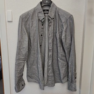 Grå skjorta från BOSS - Säljer en stilren grå skjorta från BOSS med klassisk krage och knäppning framtill. Skjortan har en bröstficka och långa ärmar med knappar vid manschetterna. Perfekt för en clean och enkel look.