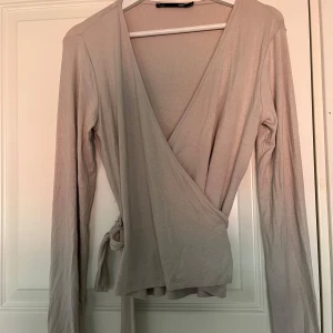Beige omlottopp från 157 - Säljer en beige omlottopp från 157 i mjukt bomullsmaterial. Toppen har långa ärmar och knytdetalj i sidan. Perfekt för lager-på-lager och enkel att styla till olika outfits. Aldrig använd 