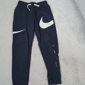 Säljer ett par mörkblåa mjukisbyxor från Nike med stora loggor - Säljer ett par mörkblåa mjukisbyxor från Nike med stora vita swoosh-loggor på benen och orange textdetaljer. Byxorna har dragsko i midjan och muddar vid bensluten. Perfekta för en sportig och avslappnad stil.