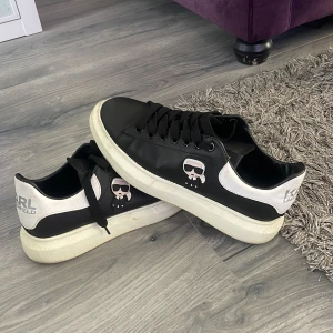 Svarta och vita sneakers från Karl Lagerfeld - Säljer ett par svarta och vita sneakers från Karl Lagerfeld med ikoniskt tryck på sidan. Skorna har snörning och en platt vit sula. Perfekta för dig som gillar stilrena och trendiga skor med en twist.