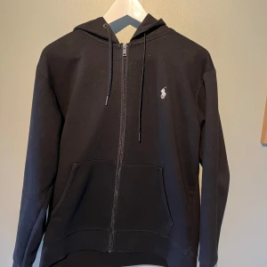Svart hoodie från Polo Ralph Lauren - Klassisk svart hoodie från Polo Ralph Lauren med dragkedja och huva. Tröjan har två fickor framtill och den ikoniska broderade loggan på bröstet. Perfekt för en avslappnad och stilren look. Säljer hoodien pga att den är för liten för mig! Kom gärna med frågor och prisförslag😁 Inga defekter😇