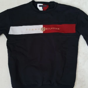 Svart sweatshirt från Tommy Hilfiger - Säljer en svart sweatshirt från Tommy Hilfiger med broderad logga och text i guld på bröstet. Tröjan har en bred rand i vitt och rött över bröstet och rund halsringning. Perfekt för dig som gillar klassisk stil med en twist.