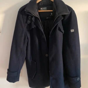 Mörkblå rock från Indigo Jeanscode - Stilren mörkblå rock från Indigo Jeanscode med klassisk krage, knappar och dragkedja framtill. Jackan har långa ärmar, fickor med lock och dekorativa detaljer vid axlar och ärmslut. En knapp är borta