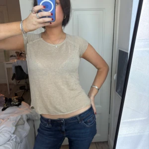 Beige topp med guldglitter från Zadig & Voltaire - Säljer en beige topp från Zadig & Voltaire med glittriga guldiga ränder. Toppen har korta ärmar, rund halsringning och dekorativa knappar på axeln. Perfekt för dig som vill ha något stilrent men ändå med lite extra detalj.