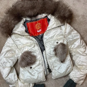 Vit pufferjacka - Säljer en vit pufferjacka från 2000-talet med stor fluffig pälskrage och pälsbollar. Jackan har dragkedja framtill och snygg brodyr på ryggen med texten 'Imperial dreams'.💞