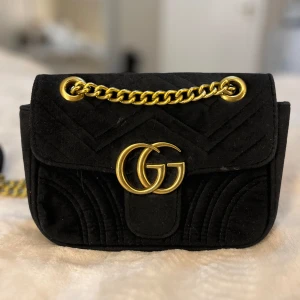 Svart sammet axelväska FEJK  - Säljer en svart axelväska i sammet med guldfärgad kedja och FEJK GG-logo framtill. Väskan har quiltade detaljer och ett hjärtmönster på baksidan. Perfekt storlek för det viktigaste och ger en lyxig känsla.