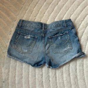 Blå jeansshorts från Just Jeans - Säljer ett par klassiska blå jeansshorts från Just Jeans. Shortsen har slitna detaljer, uppvikta benslut och dekorativa sömmar på bakfickorna. Perfekta för varma dagar och en avslappnad stil.