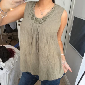 Grön ärmlös linnetopp med broderi - Säljer en grön ärmlös linnetopp med broderade detaljer och knytband vid halsen. Toppen har en avslappnad passform och är tillverkad i ett lätt, skrynklat material som ger en bohemisk känsla.