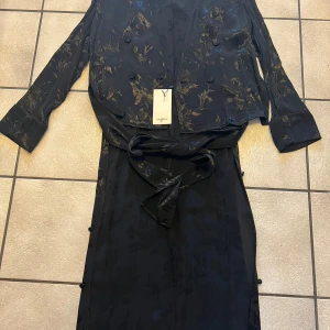 Svart kavaj med lång baksida med blommigt mönster från Zara Basic - Säljer en svart kavaj från Zara Basic med subtilt blommigt mönster i guldton. Klänningen har långa ärmar, knappar framtill och ett matchande bälte i midjan. Perfekt för dig som gillar eleganta detaljer och stilren design.