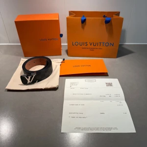 Svart LV bälte - Säljer ett exklusivt svart bälte från Louis Vuitton med klassiskt rutigt mönster och ett stilrent silverfärgat LV-spänne. Bältet är gjort i skinn och har en lyxig känsla med detaljerad sömnad och präglad logotyp på insidan.