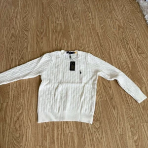 Vit kabelstickad tröja från Polo Ralph Lauren - Säljer en klassisk vit kabelstickad tröja från Polo Ralph Lauren med rund halsringning och broderad logga på bröstet. Tröjan har långa ärmar och ribbade muddar vid ärmslut och nederkant. Perfekt för en stilren look.