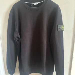 Svart sweatshirt från Stone Island - Säljer en svart sweatshirt från Stone Island med klassisk patch på vänster ärm. Tröjan har rund halsringning och är tillverkad i mjukt material. Perfekt för en stilren och avslappnad look.