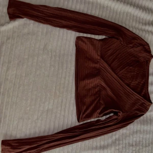 Brun omlottopp med lång ärm - Säljer en brun omlottopp med lång ärm. Toppen har en croppad passform och är tillverkad i ett glansigt, ribbat material. Perfekt för dig som gillar trendiga och stilrena plagg.