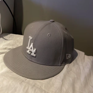 Grå LA keps från New Era 59FIFTY - Säljer en stilren grå keps från New Era 59FIFTY med LA-logga broderad framtill i vitt. Klassisk flat brim och broderade ventilationshål. Perfekt för dig som gillar feta kepsar till sommaren. Bara höra av dig om det är något😁
