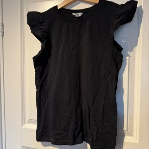 Svart topp med spetsärmar från TIKI - Svart topp från TIKI med fina spetsdetaljer på axlarna. Rund halsringning och korta ärmar med volangliknande spets. Perfekt att styla till jeans eller kjol för en trendig look.