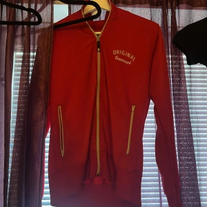 Röd track jacket från Original Greenwood - Röd track jacket från Original Greenwood med vita dragkedjor och broderad logga på bröstet. Jackan har två fickor med dragkedja och ribbade muddar vid ärmslut och nederkant. Perfekt för en sportig look.