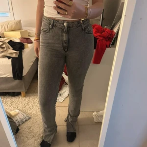 Grå raka jeans med slits - Säljer ett par grå jeans med raka ben och slits nertill. Jeansen har klassisk femficksdesign och hög midja. Perfekta för dig som gillar en enkel men trendig look.