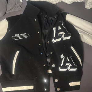 Svart och vit varsityjacka från Axel Arigato - Säljer en svart och vit varsityjacka från Axel Arigato med stora A-patchar på framsidan och broderad text på bröstet. Jackan har långa ärmar i vitt, svarta muddar och knappar framtill. Perfekt statement-plagg för streetwear-stilen.