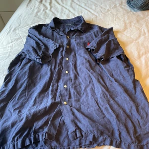 Blå kortärmad skjorta från Polo Ralph Lauren - Säljer en blå kortärmad skjorta från Polo Ralph Lauren med klassisk krage och knappar framtill. Skjortan har en röd broderad logga på bröstet och är tillverkad i bomull. Perfekt för en avslappnad och stilren look.