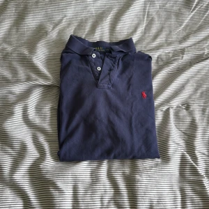 Mörkblå pikétröja från Ralph Lauren - Säljer en stilren mörkblå pikétröja från Ralph Lauren med klassisk krage och knappar. Tröjan har en broderad röd logotyp på bröstet och är kortärmad. Perfekt för en avslappnad men ändå snygg look.