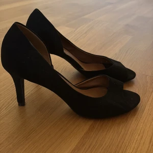 Svarta pumps med öppen tå - Säljer ett par klassiska svarta pumps med öppen tå och smal klack. Skorna har en elegant form och är tillverkade i mocka med en mjuk insida. Perfekta för dig som vill ha en stilren och tidlös sko till garderoben.