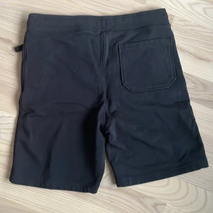 Svarta shorts med fickor - Svarta shorts med två framfickor och en bakficka. Klassisk design med normal passform och resår i midjan. Liten vit etikett nertill på ena benet. Perfekta för sommardagar.