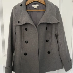 Grå kappa från H&M - Snygg grå kappa från H&M i klassisk modell med dubbelknäppning och bred krage. Jackan har långa ärmar och svarta knappar framtill. Perfekt till våren och passar till många olika outfits.