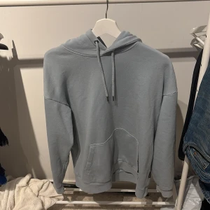 Ljusgrå hoodie med ficka - En stilren ljusgrå hoodie med huva och dragsko. Tröjan har en stor magficka framtill och ribbade muddar vid ärmslut och nederkant. Perfekt för en avslappnad look.