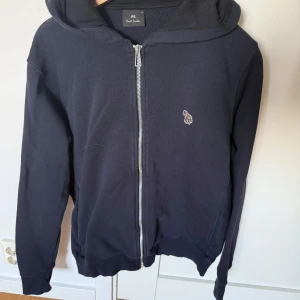 Mörkblå hoodie med dragkedja från Paul Smith - Säljer en mörkblå hoodie från Paul Smith med dragkedja framtill och liten broderad logga på bröstet. Tröjan har huva och ribbade muddar vid ärmslut och nederkant. Perfekt för en avslappnad stil.