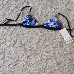 Blå och vit trekantsbikini med knyt - Säljer en snygg trekantsbikini i blått och vitt kamouflagemönster. Bikinin har svarta band och knyt i ryggen. Perfekt för stranden eller poolen! Har fortfarande prislappen kvar och kostade från början 99 men den har tyvärr en skada nu.