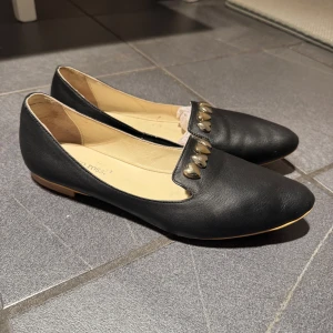 Svarta läder ballerinaskor med hjärtdetaljer  - Säljer ett par svarta ballerinaskor från Vito Rossi med spetsig tå och dekorativa hjärtan i metall framtill. Skorna har en platt sula och är tillverkade i skinn med beige innersula. Perfekta för dig som gillar unika detaljer!