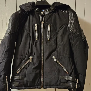 Svart dun jacka med läderdetaljer från Horspist - Säljer en dam/barn svart jacka från Horspist med coola läderdetaljer på axlar, ärmar och rygg. Jackan har flera dragkedjor framtill, huva och bältesdetaljer i midjan. Perfekt för dig som vill ha en edgy och snygg look. Jackan är väldigt liten I storleken. Finns även stor päls till luvan om så önskas.