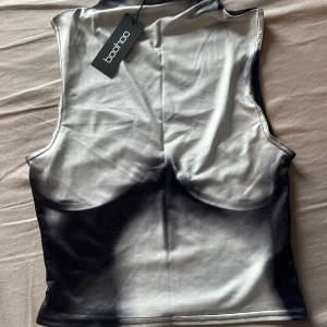 Svartvit ärmlös topp från Boohoo - Cool ärmlös topp från Boohoo i svartvitt med ett unikt tryck som ger en edgy look. Toppen har en tight passform och är tillverkad i ett mjukt syntetmaterial. Perfekt för att sticka ut i mängden.