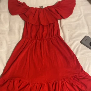 Röd offshoulder klänning från ASOS - Säljer en röd offshoulder klänning från ASOS med volang upptill och vid kjol. Klänningen har resår i midjan och är tillverkad i ett mjukt tyg som ger en bekväm passform. Perfekt för sommarens tillfällen!