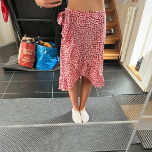 Röd blommig kjol med volang - Säljer en röd kjol med vitt blommönster och volangkant. Kjolen har knytband i midjan och är i en lätt och luftig modell som passar perfekt till varmare dagar.